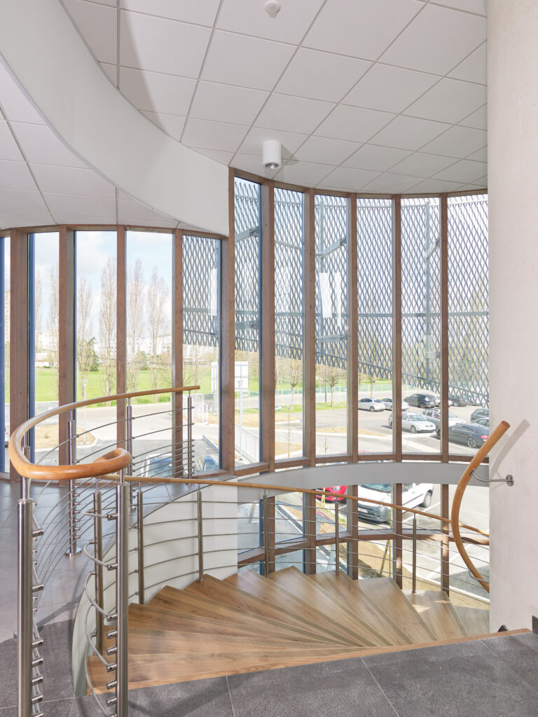 Création du show-room du Comptoir des fers à Chalon-sur-Saône par Naos Atelier d'Architecture. Vue intérieure de la cage d'escalier. Marches en bois, parois extérieures vitrées avec résille métallique